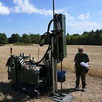 Auf einem Feld steht eine Sondierraupe, daneben steht eine Soldatin.