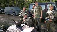 Mehrere Soldaten stehen am Waldrand und beobachten interessiert ein Szenario der Übung.