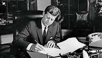 John F. Kennedy am Schreibtisch beim Unterzeichnen des Erlasses über das Verbot der Lieferung von Angriffswaffen an Kuba 