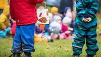 Zwei kleine Kinder stehen in Regenkleidung draußen auf dem Rasen. Im Hintergrund sind weitere spielende Kinder zu sehen