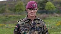 Ein Soldat steht im Feldanzug mit Barrett vor einer Wiese. Im Hintergrund ist ein leichter Hügel zu sehen