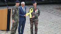 Ein Zivilist steht zwischen von zwei Soldaten vor einem Pult. Sie halten eine große gelbe Schleife.