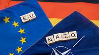 Eine Europaflagge, eine NATO Flagge und eine deutsche Flagge liegen übereinander, darüber Scrabblesteine mit EU und NATO