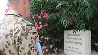 Umrandet von blumigen Büschen steht auf einer Mauer ein weißer Gedenkstein. Ein UN-Soldat steht davor und betrachtet diesen.