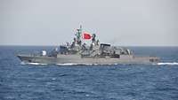 Ein graues Schiff fährt durch das Meer. Es hat die türkische Flagge gesetzt-ein weißer Sichelmond mit Stern auf rotem Grund.