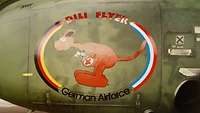 Auf einem grünen Flugzeug ist ein Bild von einem Känguru gemalt, dass eine verletzte Maus in seinem Beutel transportiert