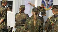 Ein Soldat zeigt mit einem Stift auf eine große Karte, die an eine Wand geheftet wurde. Davor stehen mehrere Soldaten.