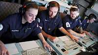 Drei Marinesoldaten in blauer Arbeitsuniform sitzen vor Computerkonsolen.