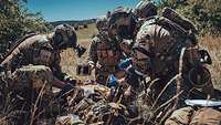 Mehrere KSK-Soldaten versorgen einen am Boden liegenden verwundeten Soldaten im Gelände.