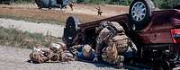Ein KSK-Soldat kniet neben einem verletzten Soldaten vor einem zerstörten Auto. Dahinter Soldaten und ein Hubschrauber am Boden.