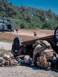 Sanitätsspezialzug Übung Ein KSK-Soldat kniet neben einem verletzten Soldaten vor einem zerstörten Auto. Dahinter Soldaten und ein Hubschrauber am Boden.