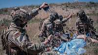 KSK-Soldaten versorgen einen verletzten Soldaten im Gelände. Ein Soldat im Vordergrund bereitet eine Infusion vor.