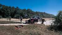 Bewaffnete KSK-Soldaten laufen auf ein zerstörtes Auto zu. Ein verletzter Soldat liegt im Gras. Hinten steht ein Hubschrauber.