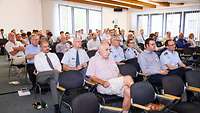 Frauen und Männer, Soldatinnen und Soldaten in einem Konferenzraum auf Stühlen.