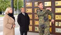 Eine Frau und zwei Männer stehen vor einer Wand mit Wappen