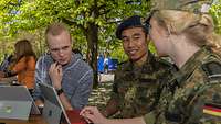Ein Zivilist, ein Soldat und eine Soldatin sitzen an einem Tisch vor Tablets und sprechen miteinander