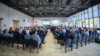 Viele Soldatinnen und Soldaten und Personen in Anzügen sitzen in einem großen Saal.