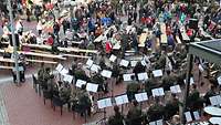 Vogelperspektive: Ein Militärorchester spielt auf einem Marktplatz, vor ihm sitzen Zivilisten.