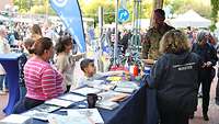 Eine Frau mit zwei Kindern steht vor einem Infostand, dahinter ein Soldat und eine Zivilistin.
