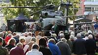 Vor einem Militärhubschrauber steht ein Pfarrer und predigt. Vor ihm sitzen Zivilisten.