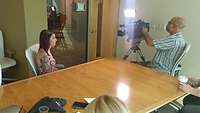 A woman sits in a meeting room and a cameraman films her with a video camera.