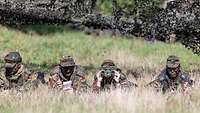 Vier Soldatinnen und Soldaten liegen im hohen Gras. Eine blickt durch ein Doppelfernglas, ein anderer schreibt etwas auf. 