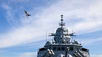 Ein Kampfjet fliegt tief über den Aufbauten eines Kriegsschiffs.