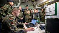 Ein Soldat sitzt an einem Schreibtisch vor einem Notebook. Mehrere Soldaten und die Verteidigungsministerin stehen daneben.