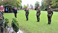 Auf einer Wiese stehen vier Soldaten nebeneinander vor Gedenkkränzen und grüßen militärisch.