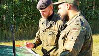 Drei Soldaten stehen an einem gepanzerten Radfahrzeug. Darüber fliegt ein Düsenjet