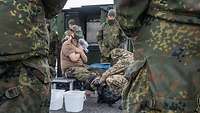 Ein Soldat mit ABC-Maske sitzt mit verschränkten Händen vor der Brust. Ihm wird ein ABC-Anzug ausgezogen.