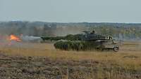 Ein getarnter Panzer steht auf einem Feld in Stellung und schießt, ein roter Feuerball entsteht.