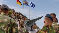 Niederländische Soldaten stehen vor drei Flaggen an einem Mast und dem HAWK-Ehrenmal.