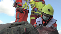 Ein Soldat liegt mit blutender Wunde am Kopf auf dem Boden, ein Sanitäter mit gelbem Schutzhelm kniet neben ihm.