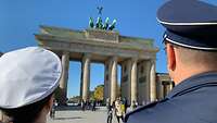 Zwischen zwei, mit dem Rücken zur Kamera stehenden Soldaten, sieht man das rege besuchte Brandenburger Tor.