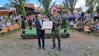Ein Zivilist im Anzug und ein Soldat in Flecktarn halten eine eingerahmte Urkunde. 