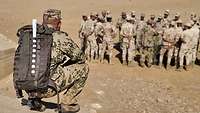 Ein deutscher Soldat mit Rucksack kniet auf einem Erdwall, in der Ferne steht eine Gruppe Soldaten.