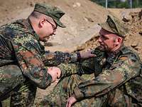 Zwei Soldaten sitzen im Gelände auf dem Boden, reichen sich dabei die Hände und sprechen miteinander