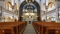 Im Betsaal einer Synagoge steht ein Mann im Anzug und spricht mit weiteren Personen auf den Sitzbänken.