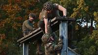 Soldaten in Flecktarn-Uniform sitzen auf einem hohen Balken; einer hilft dabei einem anderen Soldaten beim Hochklettern 