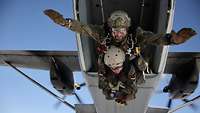 Zwei Fallschirmjäger als Tandem springen aus der geöffneten Heckklappe eines Flugzeuges.