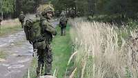 Soldat mit Waffe in der Hand im Vordergrund beobachtet den Wald abseits des Weges.