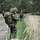 Soldat mit Waffe in der Hand im Vordergrund beobachtet den Wald abseits des Weges.