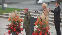 Verteidigungsministerin Lambrecht hält an einem Pult eine Rede, daneben ein Soldat und hinter ihr ein Mann mit Regenschirm