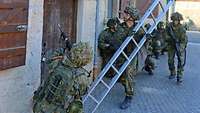 Ein Soldat stellt eine Leiter an eine Hauswand. Weitere Soldaten sichern mit ihren Gewehren.