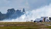 Ein Hubschrauber fliegt über einer Nebelwand; im Vordergrund steht ein Mann neben Kameras und ein paar Fahrzeugen.
