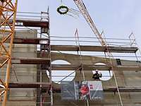 Richtspruch Pionierkaserne Auf einem Gerüst an einem Neubau steht ein Bauarbeiter mit Schutzhelm. Er hält ein Glas und einen Zettel in der Hand
