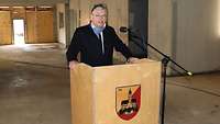 Ein Mann mit schwarzem Mantel und hellem Schal steht an einem Rednerpult in einem Rohbau und spricht