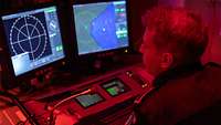Ein Marinesoldat in blauer Arbeitsuniform sitzt an einer Computerkonsole mit zwei Bildschirmen.
