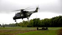 Ein Transporthubschrauber hebt von einem Feld ab. Hinter ihm warten mehrere Soldaten am Boden.
